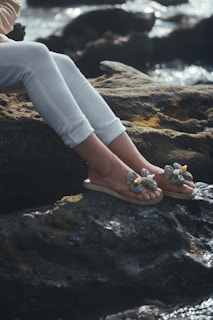 A person's legs and feet wearing decorative sandals are resting on rocky terrain by the water's edge. The person is wearing white pants, and the focus is on the detailed design of the sandals, which have floral decorations.