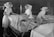 Three nurses in traditional uniforms are making beds in a hospital ward. The beds are organized in a neat row and the room has a dimly lit, monochromatic atmosphere. The nurses are focused on their tasks, arranging the white sheets with precision. In the background, dim figures of people are visible.