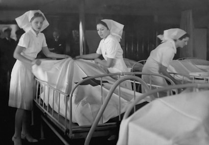 Three nurses in traditional uniforms are making beds in a hospital ward. The beds are organized in a neat row and the room has a dimly lit, monochromatic atmosphere. The nurses are focused on their tasks, arranging the white sheets with precision. In the background, dim figures of people are visible.