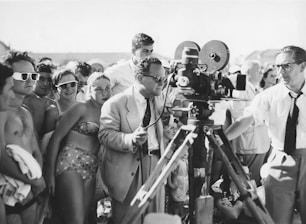 A black and white photograph of a group of people gathered around a film crew. The crew is operating a vintage film camera, with one man in a suit adjusting it. The surrounding crowd appears to be a mix of casually dressed individuals, possibly at a beach or outdoor event, with some wearing swimwear and sunglasses.
