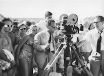 A black and white photograph of a group of people gathered around a film crew. The crew is operating a vintage film camera, with one man in a suit adjusting it. The surrounding crowd appears to be a mix of casually dressed individuals, possibly at a beach or outdoor event, with some wearing swimwear and sunglasses.