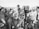 A black and white photograph of a group of people gathered around a film crew. The crew is operating a vintage film camera, with one man in a suit adjusting it. The surrounding crowd appears to be a mix of casually dressed individuals, possibly at a beach or outdoor event, with some wearing swimwear and sunglasses.