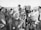 A black and white photograph of a group of people gathered around a film crew. The crew is operating a vintage film camera, with one man in a suit adjusting it. The surrounding crowd appears to be a mix of casually dressed individuals, possibly at a beach or outdoor event, with some wearing swimwear and sunglasses.