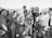 A black and white photograph of a group of people gathered around a film crew. The crew is operating a vintage film camera, with one man in a suit adjusting it. The surrounding crowd appears to be a mix of casually dressed individuals, possibly at a beach or outdoor event, with some wearing swimwear and sunglasses.