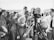 A black and white photograph of a group of people gathered around a film crew. The crew is operating a vintage film camera, with one man in a suit adjusting it. The surrounding crowd appears to be a mix of casually dressed individuals, possibly at a beach or outdoor event, with some wearing swimwear and sunglasses.