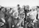 A black and white photograph of a group of people gathered around a film crew. The crew is operating a vintage film camera, with one man in a suit adjusting it. The surrounding crowd appears to be a mix of casually dressed individuals, possibly at a beach or outdoor event, with some wearing swimwear and sunglasses.