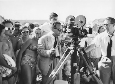 A black and white photograph of a group of people gathered around a film crew. The crew is operating a vintage film camera, with one man in a suit adjusting it. The surrounding crowd appears to be a mix of casually dressed individuals, possibly at a beach or outdoor event, with some wearing swimwear and sunglasses.