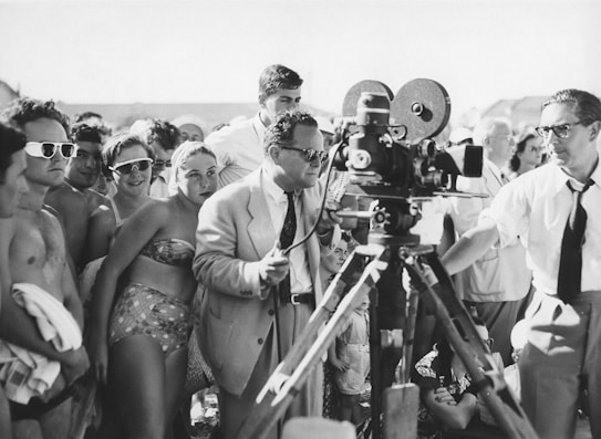 A black and white photograph of a group of people gathered around a film crew. The crew is operating a vintage film camera, with one man in a suit adjusting it. The surrounding crowd appears to be a mix of casually dressed individuals, possibly at a beach or outdoor event, with some wearing swimwear and sunglasses.