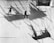 Two workers are suspended on wooden planks, painting or maintaining the surface of a large metal structure. Long shadows are cast on the wall, creating striking geometric patterns.