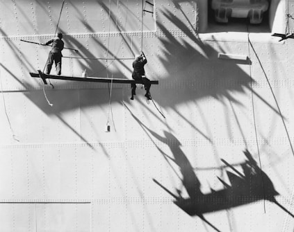 Two workers are suspended on wooden planks, painting or maintaining the surface of a large metal structure. Long shadows are cast on the wall, creating striking geometric patterns.