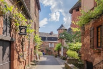 A picturesque cobblestone street is flanked by charming, old stone buildings with rustic architecture. Vines and greenery adorn the walls and windows, adding a touch of nature. The warm tones of the stone contrast with the vibrant green plants and the bright blue sky dotted with clouds. A few signs and planters decorate the street, hinting at a quaint village atmosphere.