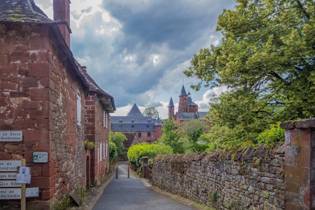 A narrow cobblestone street leads through a quaint village featuring rustic brick buildings with slate roofs. Lush greenery lines the path, while a large tree provides partial shade. In the distance, a historic church with tall steeples adds a medieval charm against a backdrop of dramatic, overcast skies.