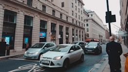 A busy city street lined with historic stone buildings. Several cars are on the road, including a silver hatchback and a black SUV. A person is walking along the sidewalk. Retail and bank signs, such as Metro Bank, are visible among the shopfronts.