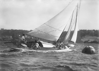 A sailboat with multiple sails is navigating through the water. Several individuals are aboard, managing the ropes and sails. The shoreline in the background is slightly blurred, suggesting movement, and a buoy is visible in the water.
