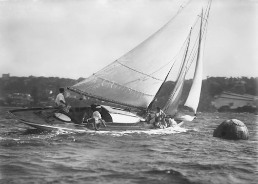 A sailboat with multiple sails is navigating through the water. Several individuals are aboard, managing the ropes and sails. The shoreline in the background is slightly blurred, suggesting movement, and a buoy is visible in the water.
