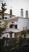 A residential building with white walls and dark window shutters. There is laundry, including a bright orange piece, hanging out to dry on a clothesline under an overhang. The roof features several chimneys, and a weather vane is perched on one of them. In the foreground, blurred foliage frames the scene, adding a natural touch.