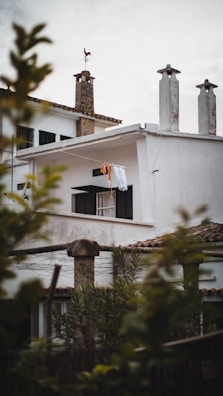 A residential building with white walls and dark window shutters. There is laundry, including a bright orange piece, hanging out to dry on a clothesline under an overhang. The roof features several chimneys, and a weather vane is perched on one of them. In the foreground, blurred foliage frames the scene, adding a natural touch.