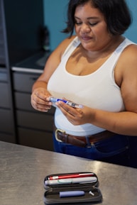 A woman is standing in a kitchen, holding a syringe and looking at it attentively. She is wearing a white tank top and blue jeans. On the metallic countertop in front of her, there is an open case containing insulin pens and a cap.