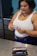 A woman is standing in a kitchen, holding a syringe and looking at it attentively. She is wearing a white tank top and blue jeans. On the metallic countertop in front of her, there is an open case containing insulin pens and a cap.
