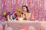A young woman with long dark hair and a tiara sits at a pink checkered table, surrounded by colorful balloons and a birthday cake. Behind her, iridescent streamers and large foil balloons form a festive backdrop. A pair of high-heeled shoes and a party hat reading 'Happy Birthday' add to the celebratory atmosphere.