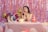A young woman with long dark hair and a tiara sits at a pink checkered table, surrounded by colorful balloons and a birthday cake. Behind her, iridescent streamers and large foil balloons form a festive backdrop. A pair of high-heeled shoes and a party hat reading 'Happy Birthday' add to the celebratory atmosphere.
