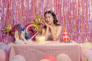 A young woman with long dark hair and a tiara sits at a pink checkered table, surrounded by colorful balloons and a birthday cake. Behind her, iridescent streamers and large foil balloons form a festive backdrop. A pair of high-heeled shoes and a party hat reading 'Happy Birthday' add to the celebratory atmosphere.