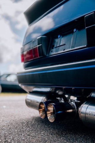 A close-up view of the rear part of a blue car, highlighting its exhaust system and license plate area. The car has dual exhaust pipes with a metallic finish, and the background is slightly blurred showing another car and the sky with clouds.