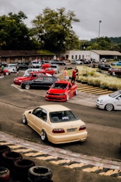 A group of modified cars parked in a lot with people gathered around them. The scene includes several vibrant cars, notably a red car in the center, surrounded by other cars of different colors. A building and trees are visible in the background, with cloudy skies above.