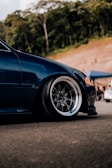 A close-up view of the front end of a dark blue car with polished alloy wheels. The setting appears to be an outdoor location with trees in the background. The car is parked on asphalt, and there is a blue tent and another vehicle in the distance.