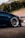 A close-up view of the front end of a dark blue car with polished alloy wheels. The setting appears to be an outdoor location with trees in the background. The car is parked on asphalt, and there is a blue tent and another vehicle in the distance.