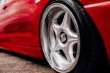A close-up of a shiny, silver alloy wheel on a red sports car. The tire is detailed with tread patterns and branding visible on the side. The vibrant red body paint reflects light, highlighting the car’s sleek design and curves.