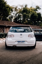 A white Volkswagen car with lowered suspension, positioned on a paved surface near a rustic building and surrounded by greenery. The vehicle has a customized license plate from Brazil and a decal on the rear windshield.