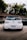 A white Volkswagen car with lowered suspension, positioned on a paved surface near a rustic building and surrounded by greenery. The vehicle has a customized license plate from Brazil and a decal on the rear windshield.