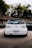 A white Volkswagen car with lowered suspension, positioned on a paved surface near a rustic building and surrounded by greenery. The vehicle has a customized license plate from Brazil and a decal on the rear windshield.