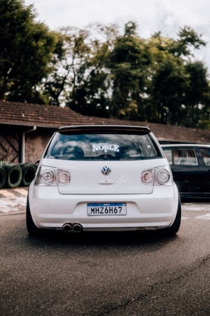 A white Volkswagen car with lowered suspension, positioned on a paved surface near a rustic building and surrounded by greenery. The vehicle has a customized license plate from Brazil and a decal on the rear windshield.