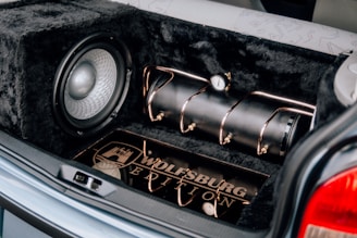 A car trunk features a custom sound system with a large subwoofer, a black compressed air tank secured with copper-colored tubes, and a custom Wolfsburg Edition plate. The interior is lined with a plush, furry black material.