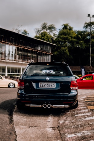 A black Volkswagen car is parked on the street with a sticker labeled 'Noble' on the rear window. The license plate reads AAY-0140. The background features a modern building with large windows and some greenery.