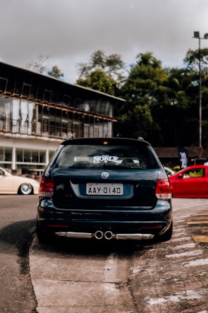 A black Volkswagen car is parked on the street with a sticker labeled 'Noble' on the rear window. The license plate reads AAY-0140. The background features a modern building with large windows and some greenery.