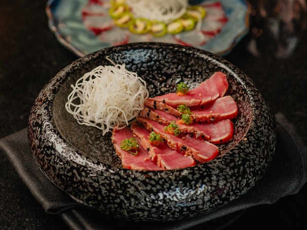 A beautifully presented dish featuring slices of seared tuna garnished with herbs, arranged alongside a portion of thinly sliced white radish. The food is placed in a textured black bowl, with another dish visible in the background blurred out.