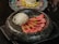 A beautifully presented dish featuring slices of seared tuna garnished with herbs, arranged alongside a portion of thinly sliced white radish. The food is placed in a textured black bowl, with another dish visible in the background blurred out.