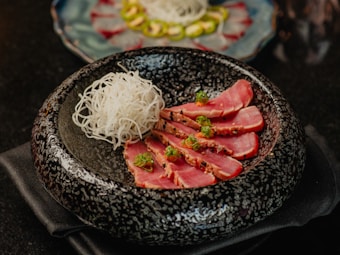 A beautifully presented dish featuring slices of seared tuna garnished with herbs, arranged alongside a portion of thinly sliced white radish. The food is placed in a textured black bowl, with another dish visible in the background blurred out.