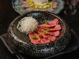 A beautifully presented dish featuring slices of seared tuna garnished with herbs, arranged alongside a portion of thinly sliced white radish. The food is placed in a textured black bowl, with another dish visible in the background blurred out.