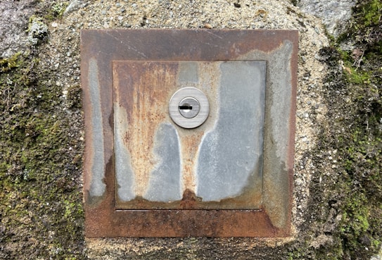 A rusty, square metal plate with a central lock embedded in a concrete wall. The metal surface is heavily weathered, showing patches of rust and discoloration. Surrounding the plate is rough concrete, partially covered with moss and small plants.