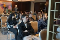 A group of people is seated in a conference or meeting room. The participants appear attentive and are dressed in casual to semi-formal attire. The room has a modern design with an abstract painting on the wall and soft lighting from overhead fixtures.