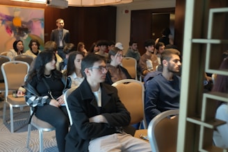 A group of people is seated in a conference or meeting room. The participants appear attentive and are dressed in casual to semi-formal attire. The room has a modern design with an abstract painting on the wall and soft lighting from overhead fixtures.