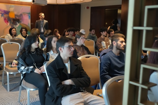 A group of people is seated in a conference or meeting room. The participants appear attentive and are dressed in casual to semi-formal attire. The room has a modern design with an abstract painting on the wall and soft lighting from overhead fixtures.