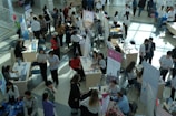 Numerous individuals are gathered in a spacious, well-lit hall, interacting around various booths and tables. Some tables are adorned with informational posters and brochures. People are engaged in discussions, examining flyers, and working on laptops. The setting appears to be a university or career fair, with banners and promotional materials visible in the background.