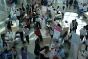 Numerous individuals are gathered in a spacious, well-lit hall, interacting around various booths and tables. Some tables are adorned with informational posters and brochures. People are engaged in discussions, examining flyers, and working on laptops. The setting appears to be a university or career fair, with banners and promotional materials visible in the background.