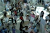 Numerous individuals are gathered in a spacious, well-lit hall, interacting around various booths and tables. Some tables are adorned with informational posters and brochures. People are engaged in discussions, examining flyers, and working on laptops. The setting appears to be a university or career fair, with banners and promotional materials visible in the background.