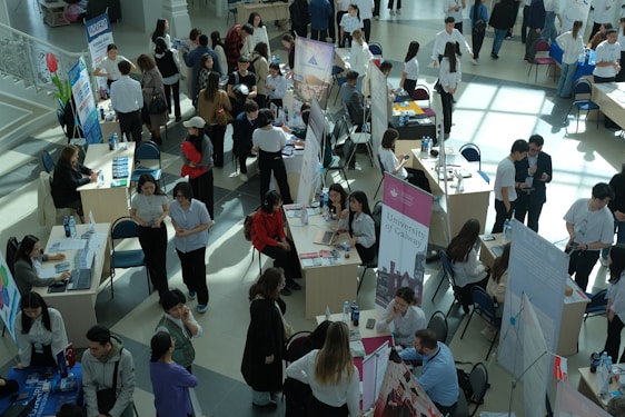 Numerous individuals are gathered in a spacious, well-lit hall, interacting around various booths and tables. Some tables are adorned with informational posters and brochures. People are engaged in discussions, examining flyers, and working on laptops. The setting appears to be a university or career fair, with banners and promotional materials visible in the background.